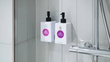 Deux flacons blancs avec des pompes noires, dont l’un est étiqueté « GO DOUBLE HAIR & BODY » et l’autre « GO SMOOTH CONDITIONER », sont fixés sur un mur carrelé dans une salle de bain.