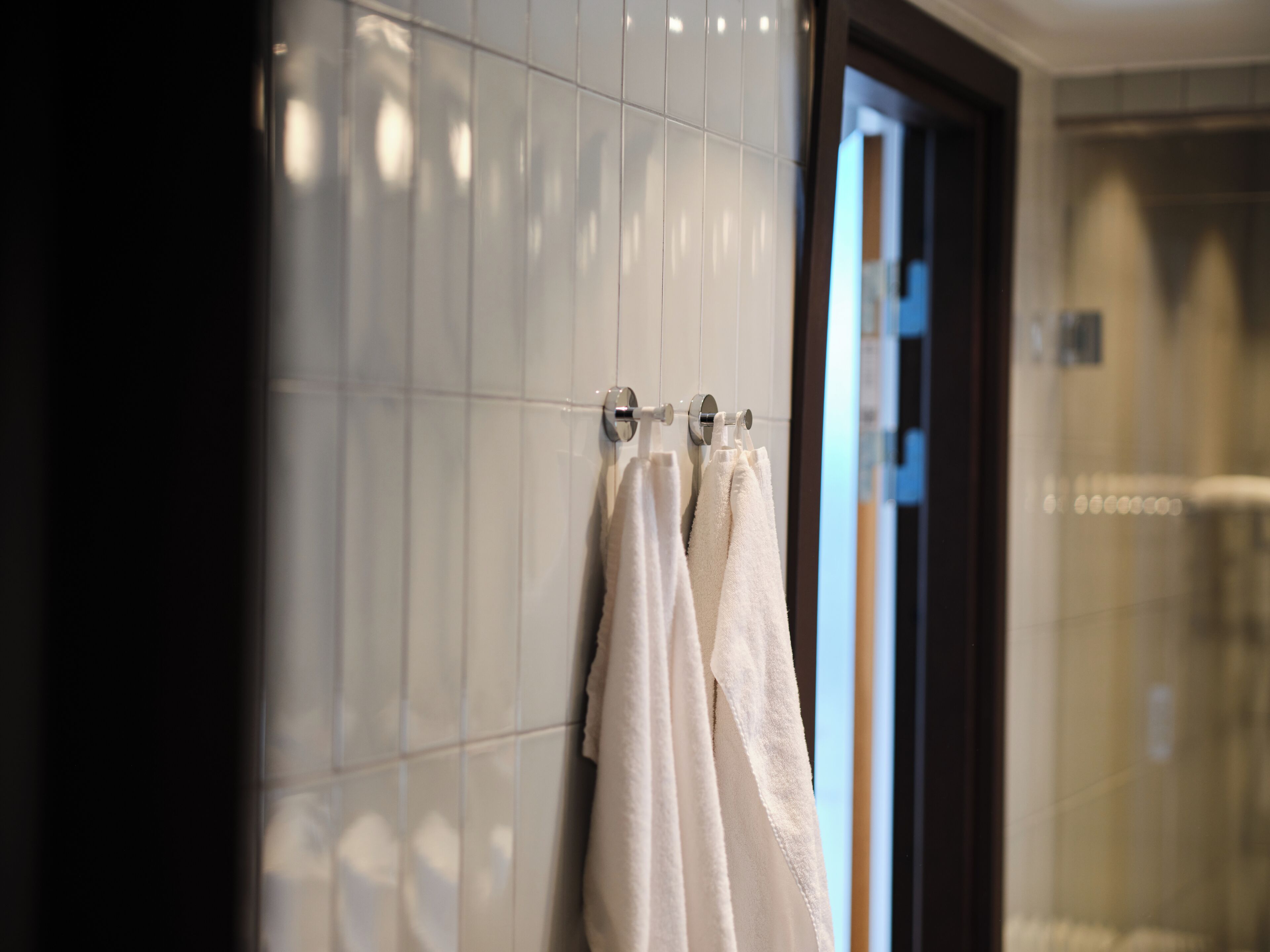 A bathroom with white tiled walls, a glass shower enclosure, and two white towels hanging on a metal towel rack.