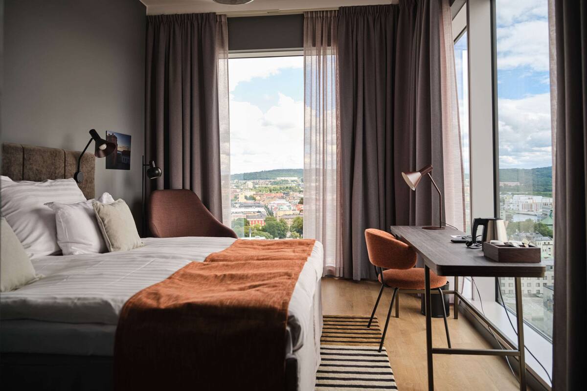 Une chambre d’hôtel équipée d’un lit, d’un bureau, d’une chaise et d’une grande fenêtre donnant sur la ville.