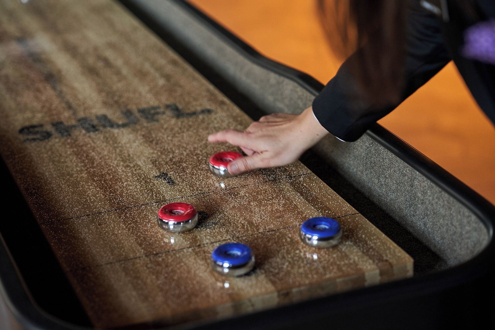 Una mesa de shuffleboard con tres discos, uno rojo y dos azules, y una mano señalando el disco rojo.