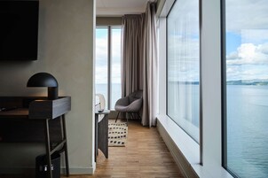 Habitación de hotel moderna con un ventanal grande, un escritorio con lámpara, una silla y vista al mar.