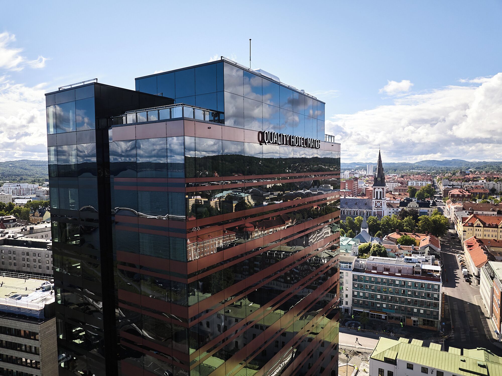 A modern hotel with a glass facade reflecting the surrounding cityscape, a prominent clock tower, and a view of the city from its rooftop.