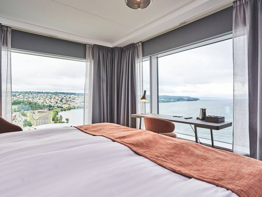 Habitación de hotel con una cama grande, un escritorio con lámpara y vistas a un río y pueblo.