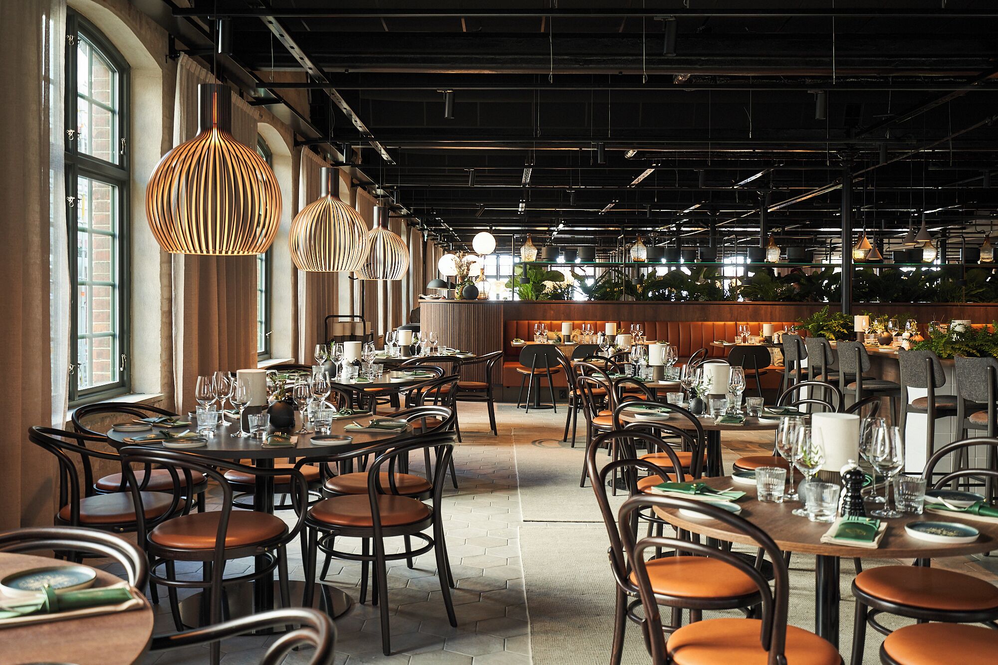 Interior de restaurante con varias mesas y sillas, ventanas amplias y lámparas colgantes.