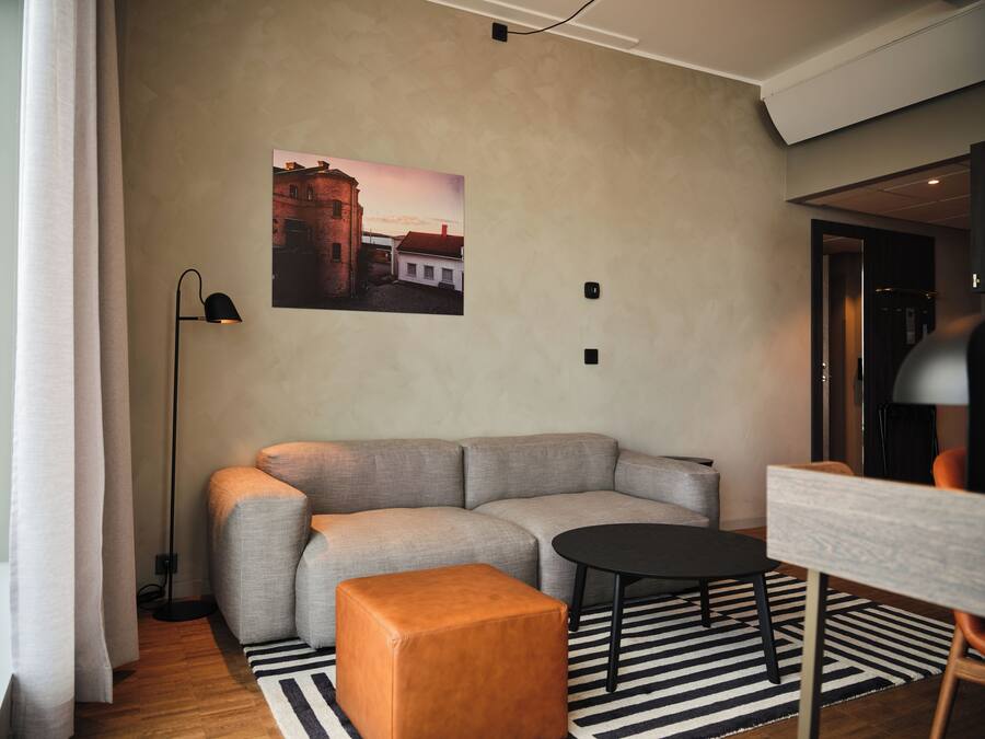 A modern living room with a grey sectional sofa, a round black coffee table, and a wooden ottoman. A black floor lamp is positioned to the left, and a black and white striped rug covers the floor.