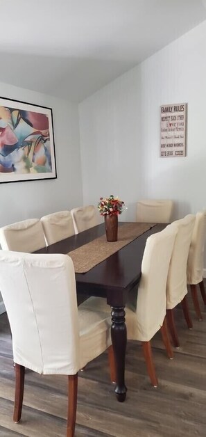 Une salle à manger avec une table en bois foncé et des chaises de couleur crème, un centre de table floral et un panneau affichant les règles de la famille, accroché au mur.