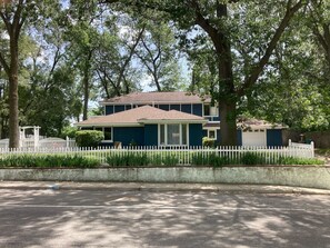 Une maison à deux étages, avec une clôture en bois peinte en blanc, entourée d’arbres.