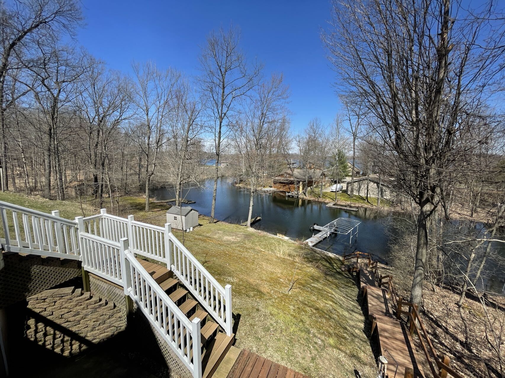 A wooden deck with a white railing overlooking a river, surrounded by leafless trees and a clear blue sky.