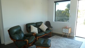 A modern living room with two green leather chairs, a small wooden side table, and a large window offering a view of trees. - Situated across the road from the beach (Bridport)