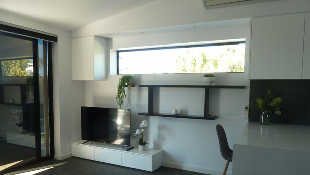 A modern living room with a flat-screen TV, a coffee table, and a plant. There is a window with a view of trees and a shelf with decorative items. - Situated across the road from the beach (Bridport)