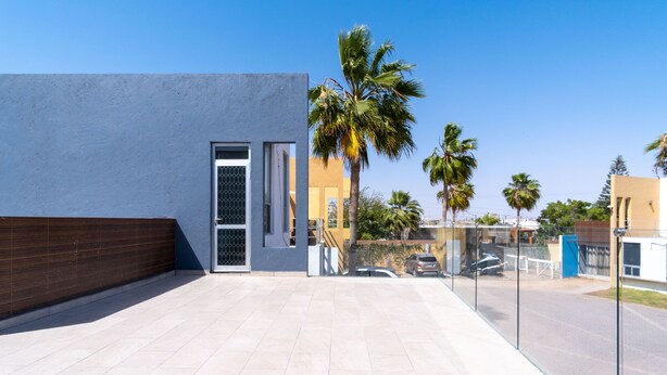 Ein modernes Gebäude mit Glastür, einer gefliesten Terrasse und Palmen im Hintergrund.