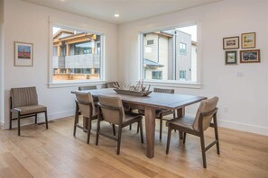 A dining room with a wooden table and six chairs, large windows with views of neighboring houses, and framed pictures on the wall.