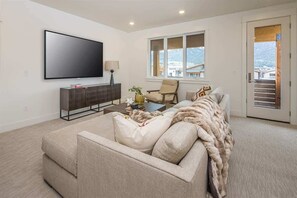 A modern living room with a flat-screen TV, a sofa, a coffee table, and a chair. There is a window with a view of a mountain.