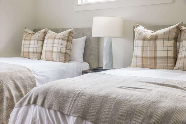 A hotel room with two beds, each with a plaid-patterned pillow and a white blanket. There is a bedside table with a lamp between the beds.
