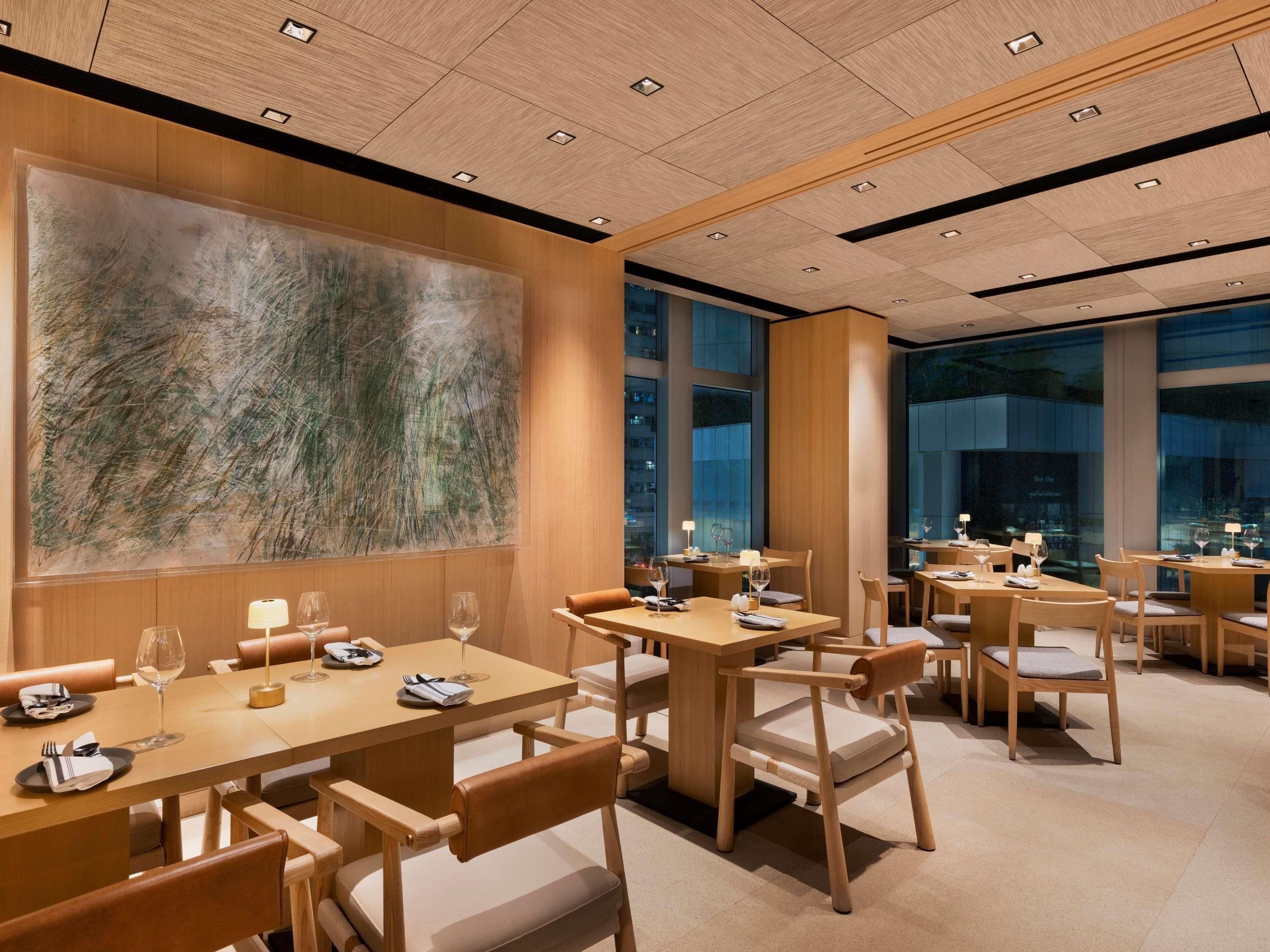 A modern restaurant interior with wooden tables and chairs, large windows, and a large abstract painting on the wall.