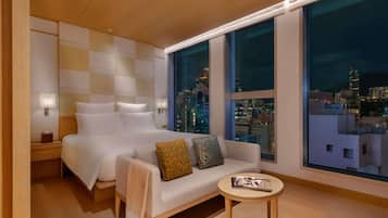 A modern hotel room with a large bed, a sofa, a small table, and a city view through large windows.
