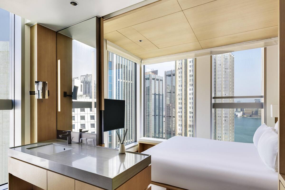 A modern hotel room with a large bed, a bathroom with a sink and mirror, and a city view through large windows.