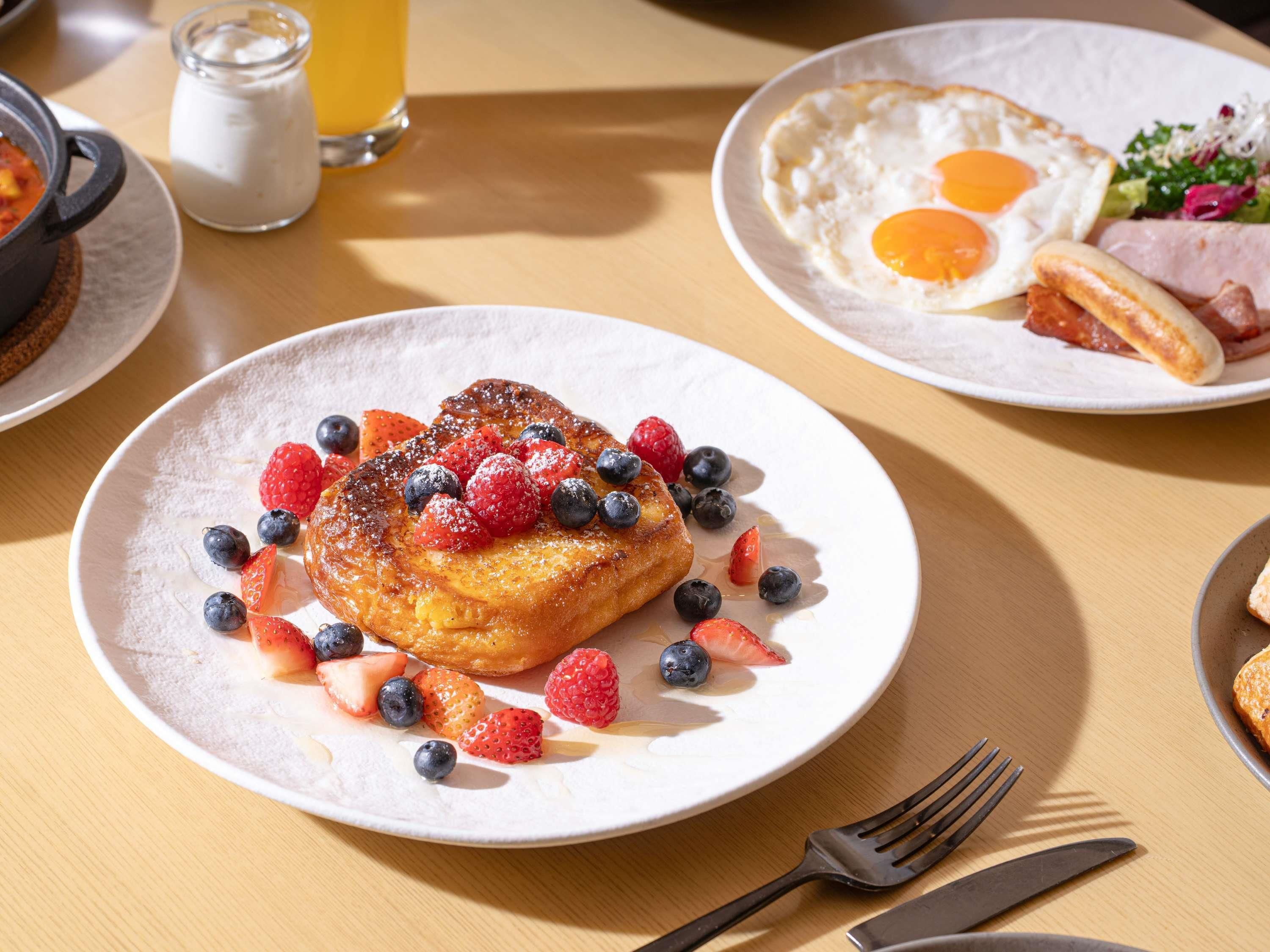 Une assiette de petit-déjeuner comprenant des crêpes, des fraises, des myrtilles, ainsi qu’une portion d’œufs brouillés et de jambon.
