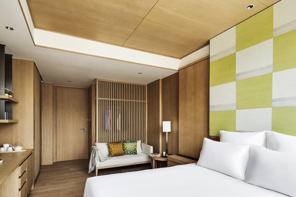 A hotel room with a bed, bedside table, lamp, and a seating area with a sofa and pillows.