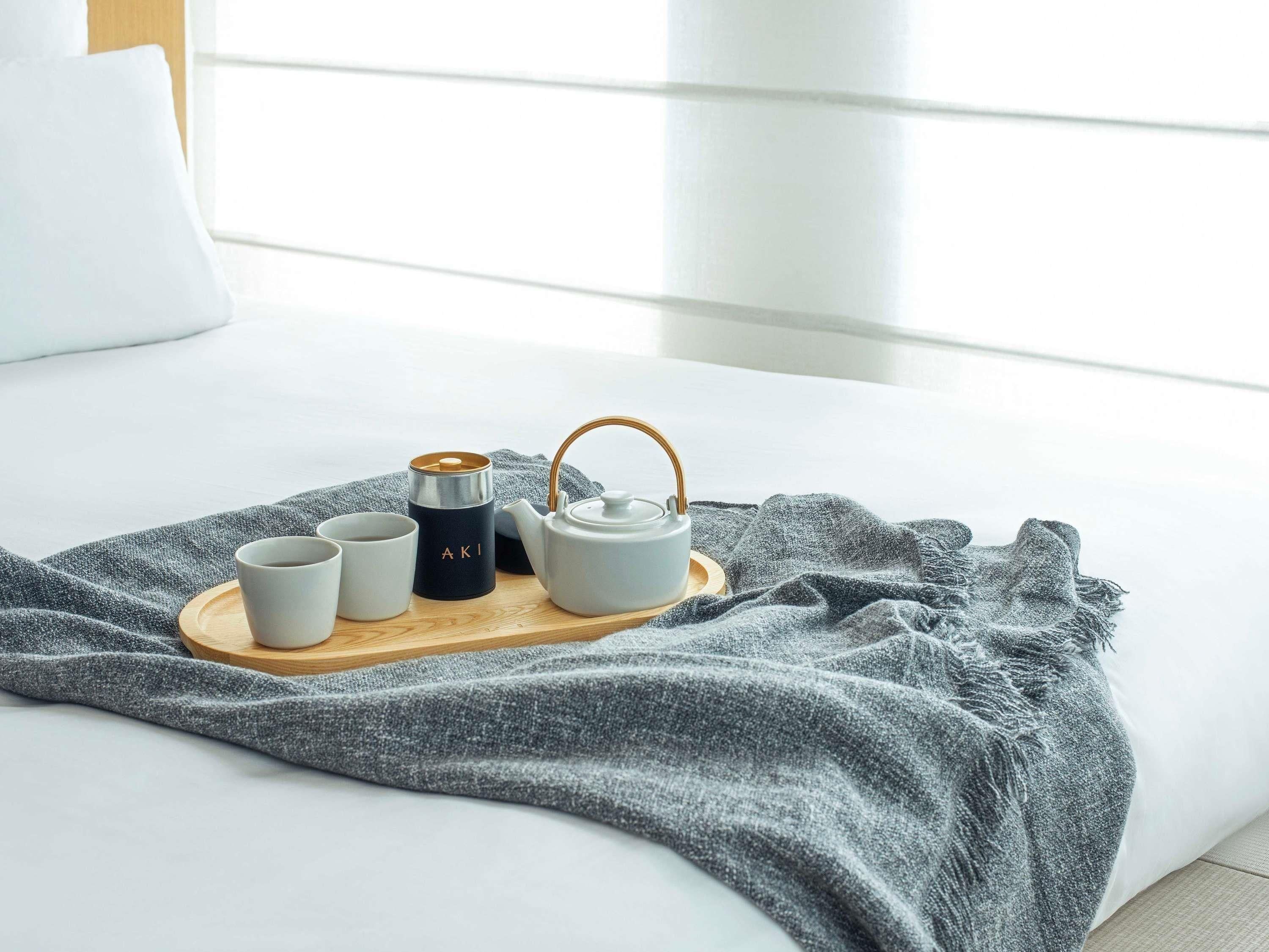 Een netjes opgemaakte ontbijtpresentatie op een bed met een grijze deken, inclusief een theepot, kopjes en een thermosfles.