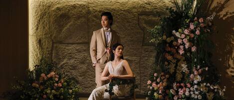 A man and woman in formal attire seated in a room with a stone wall and floral arrangements.
