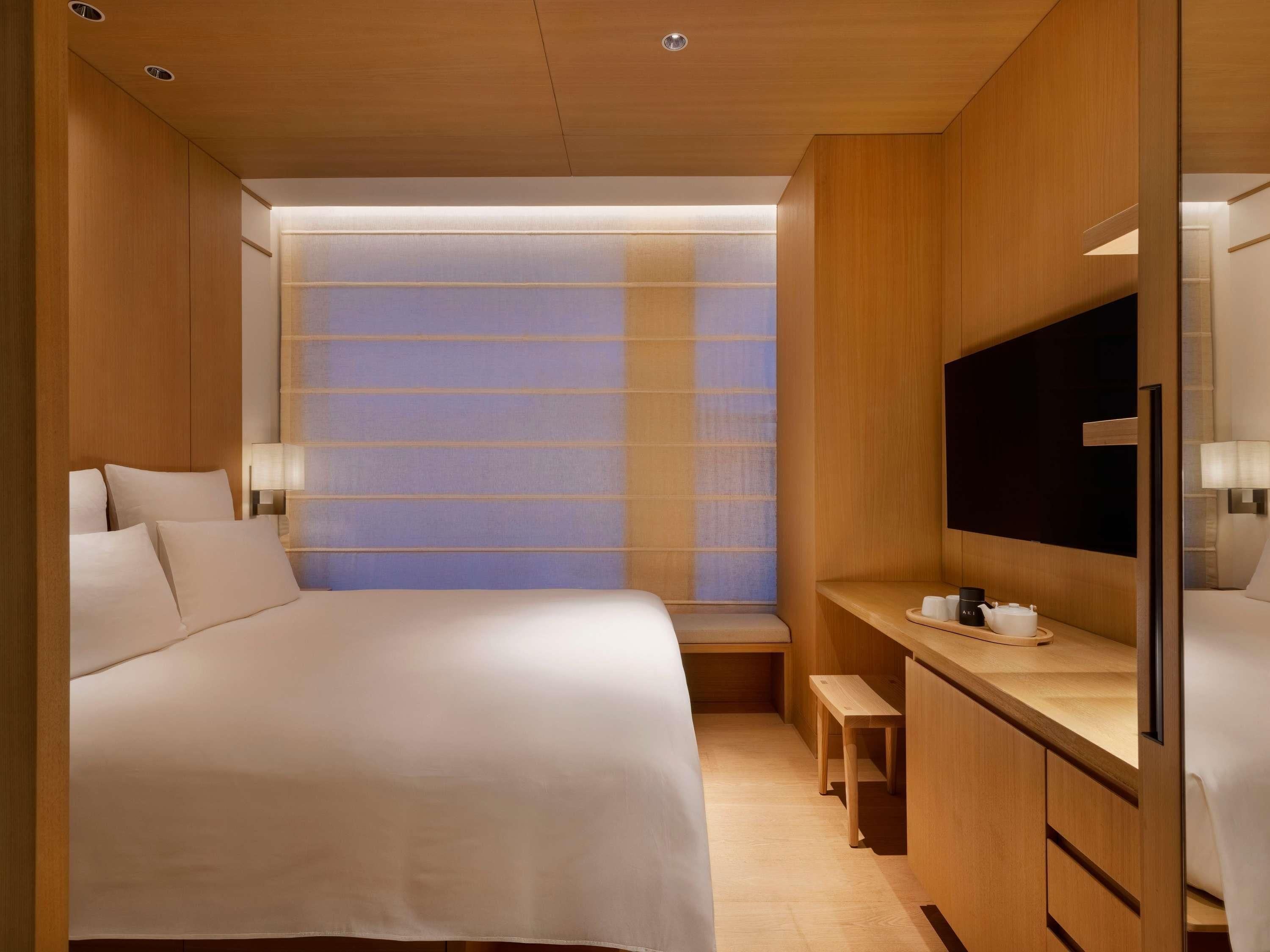 Ein modernes Hotelzimmer mit Bett, Nachttisch, Fernseher und einem Fenster mit Jalousien.