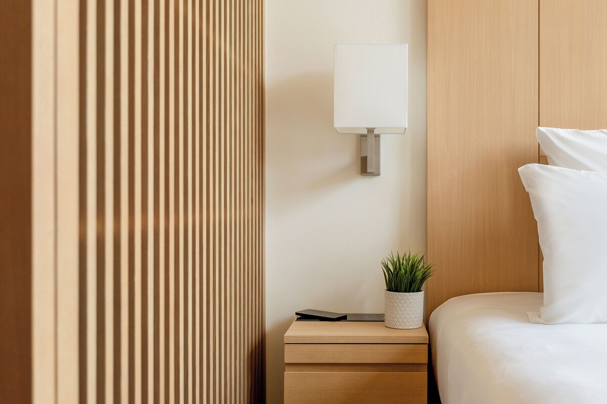 Une chambre d’hôtel bien rangée, avec une table de chevet en bois, une lampe et une plante en pot.