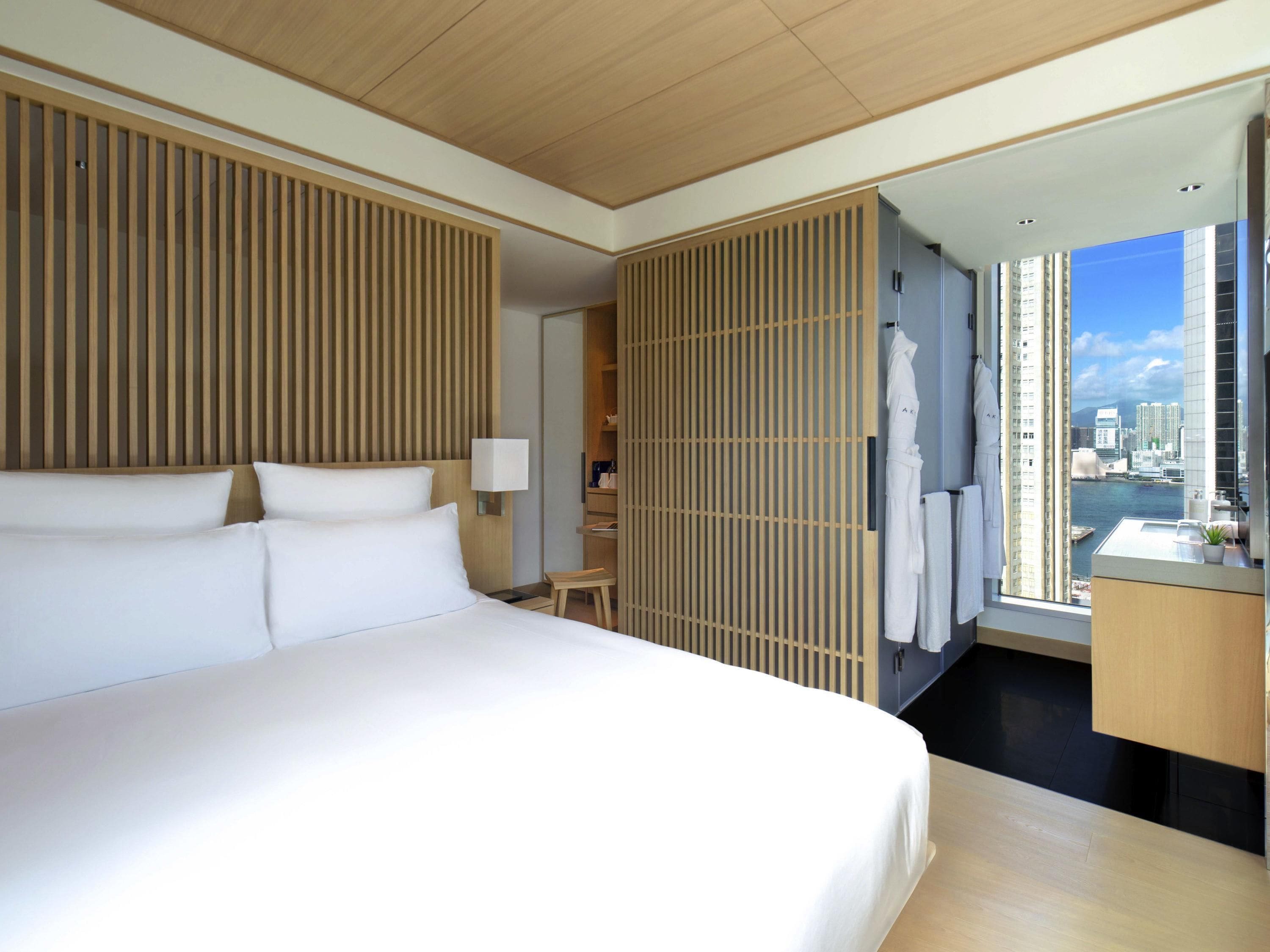 A hotel room with a large bed, a wooden partition, a small table, a chair, a desk with a mirror, and a view of a cityscape.