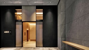 A modern hotel entrance with a dark facade, a visible elevator, and a wooden bench outside.