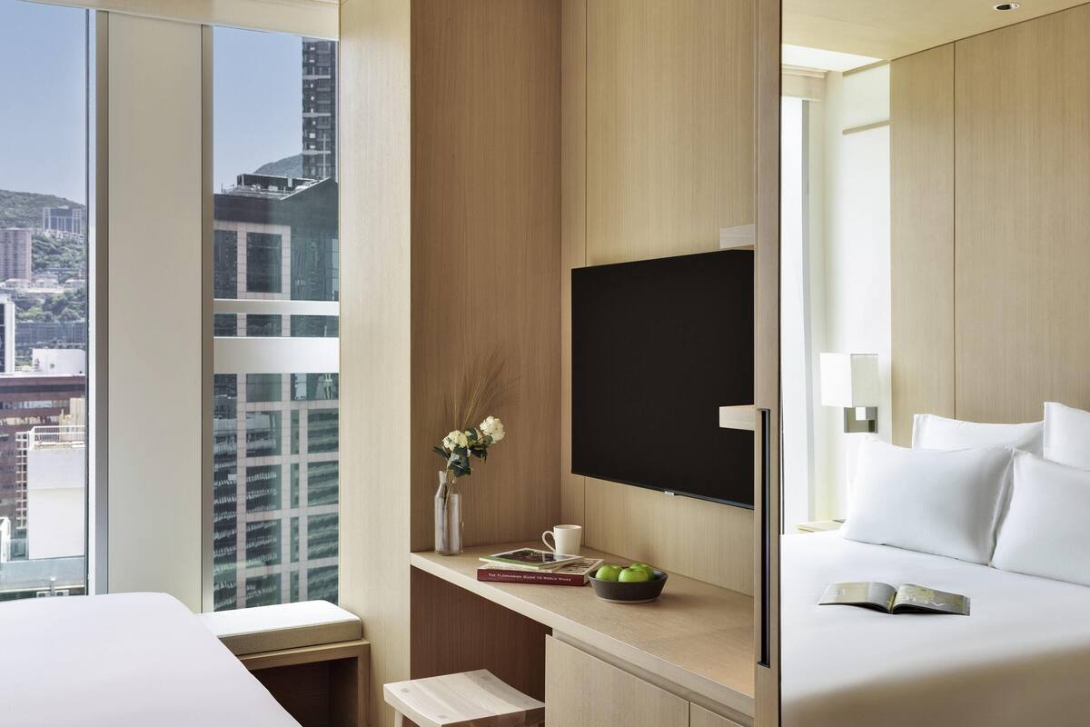 A modern hotel room with a large window, a flat-screen TV, a bedside table with a book and a bowl of fruit, and a bed with white bedding.
