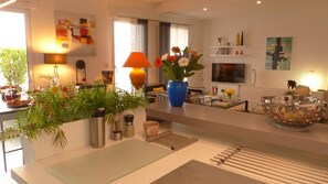 Een moderne keuken met een centraal eiland, een eetgedeelte met een glazen tafel, en een woonruimte met een televisie en kunstwerken.