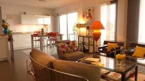Een moderne woonkamer met een glazen salontafel, een bank en een stoel met een kleurrijk kussen. Er is een keuken met witte kasten en een eettafel met rode stoelen.