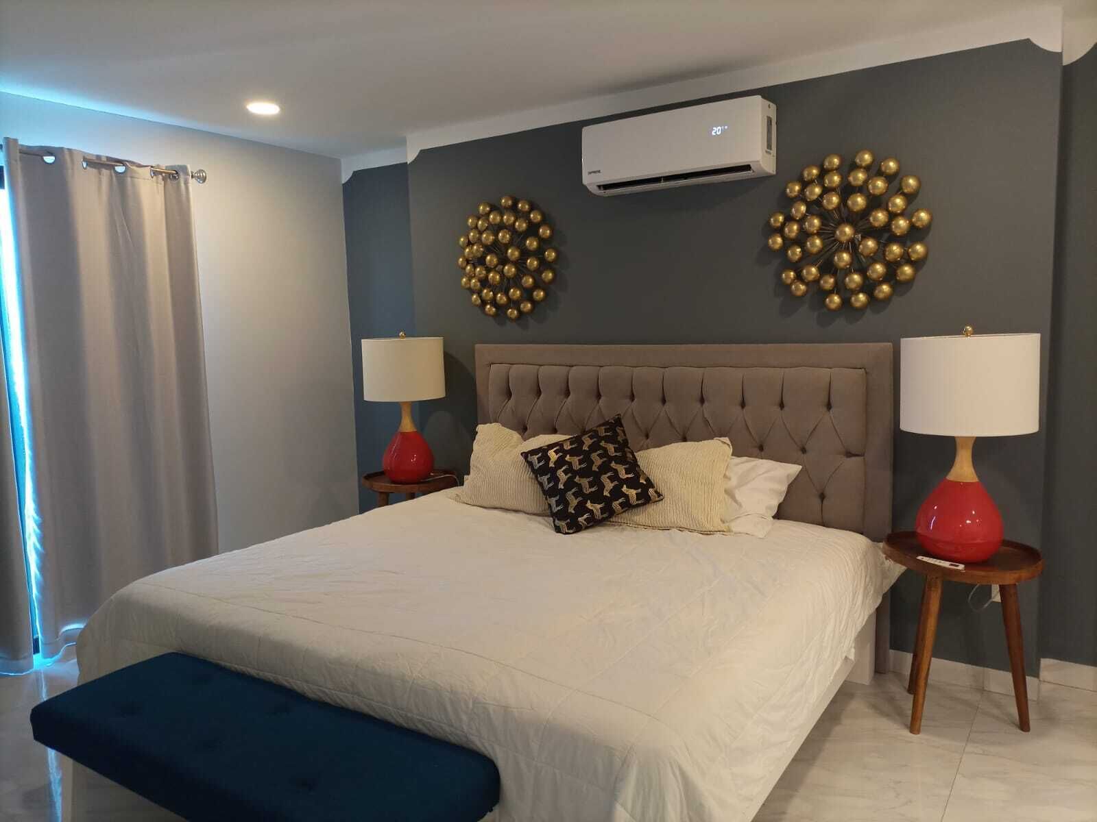 Un dormitorio con una cama grande, dos mesitas de noche con lámparas, un banco y decoración en la pared.