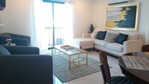 Ein Wohnzimmer mit einer Couch, Sesseln, einem Glastisch und einem Esstisch mit Stühlen. Es gibt ein großes Fenster mit Vorhängen, eine Klimaanlage und ein Bild an der Wand.
