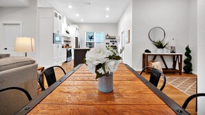 Un coin repas avec une table en bois, des chaises et un vase de fleurs. Un canapé et une lampe sont visibles au premier plan. La pièce a des murs blancs et une cuisine à l’arrière-plan.