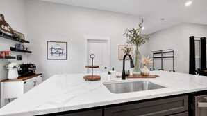Une cuisine moderne avec un plan de travail en marbre blanc, un évier en acier inoxydable et des meubles de couleur sombre. On y trouve également des éléments décoratifs, comme un vase avec des fleurs et un tableau encadré accroché au mur.