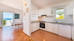 A modern kitchen with white cabinets, a built-in oven, and a dishwasher. There is a dining area with a table and chairs, and a large window offering a view of the sea. - Stunning 3-bedroom Apartment in Gradac (Gradac)