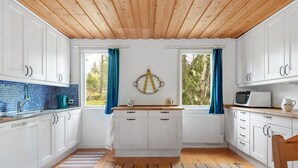 Eine Küche mit weißen Schränken, einer Holzbalkendecke, einem Fenster mit blauen Vorhängen, einer Mikrowelle und einem Wasserkocher.