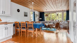 Eine geräumige Küche mit Holzausstattung, ein Essbereich mit Tisch und Stühlen sowie ein Blick ins Freie durch große Fenster.