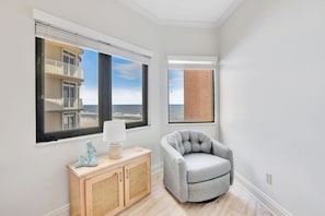 Une pièce avec vue sur un immeuble et la mer, un fauteuil gris, un meuble en bois et une lampe.