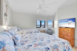 A bedroom with a large bed, a ceiling fan, a TV, and a view of the ocean through the windows.