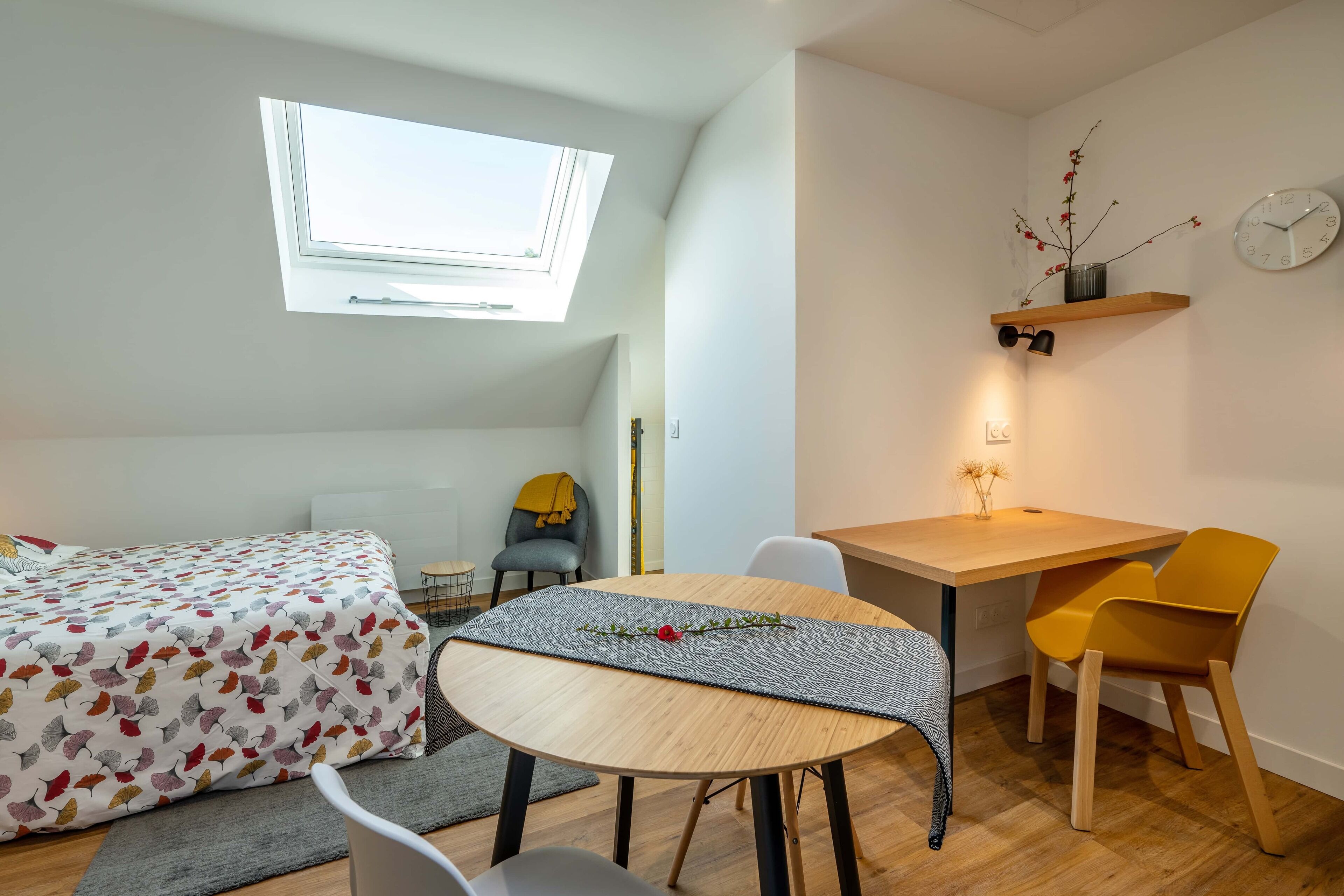 A compact room with a bed, a small table with chairs, a skylight, a clock, and a shelf with decorative items.