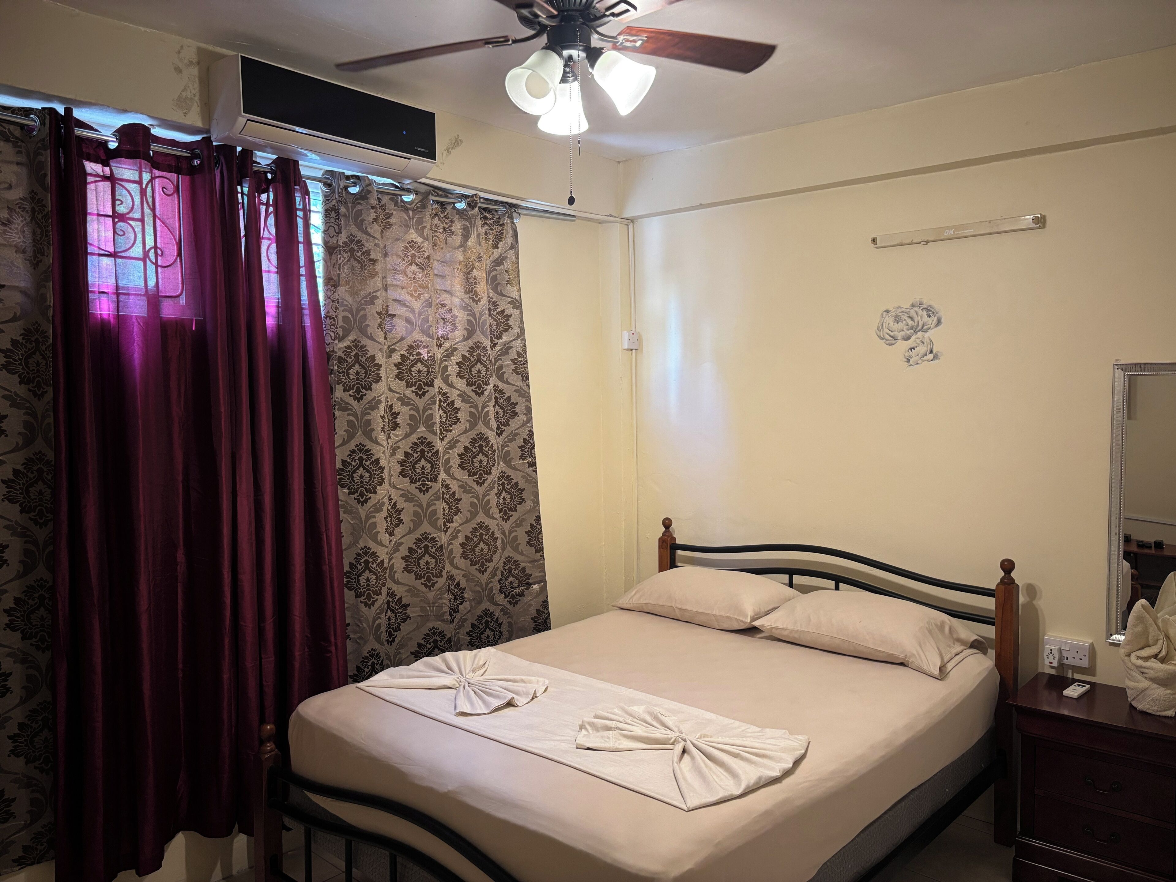 A hotel room with a bed, a ceiling fan, a wall-mounted air conditioner, and a bedside table with a lamp.