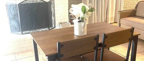 Un coin repas avec une table en bois, des chaises noires, un vase blanc orné de fleurs, un téléviseur à écran plat fixé à un mur en briques et une cheminée.