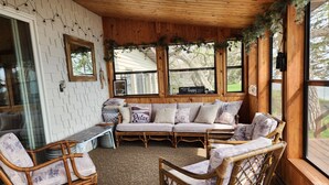 A cozy room with a wooden ceiling, a cushioned sofa, wicker chairs, and a window with a view of trees. - Beautiful, family friendly Lake home w/sunroom and 100ft of sandy beach! (Henning)