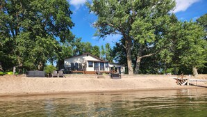 A lakeside cabin with a deck, surrounded by trees and a sandy shoreline. - Beautiful, family friendly Lake home w/sunroom and 100ft of sandy beach! (Henning)