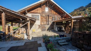 A rustic wooden cabin with a sloped roof, a stone staircase leading to a wooden door, and a covered patio area with potted plants and firewood stacks.