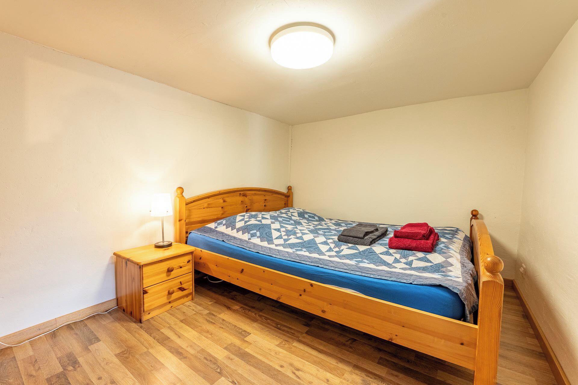 A small, single-bed room with a wooden headboard, a bedside table with a lamp, and a wooden floor.