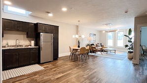 A modern kitchen with dark cabinets, a stainless steel refrigerator, and a dining area with a wooden table and chairs. - Historic & Modern! Walk to Restaurants, Moccasin Springs + Huge Suite w/ Kitchen (Hot Springs)