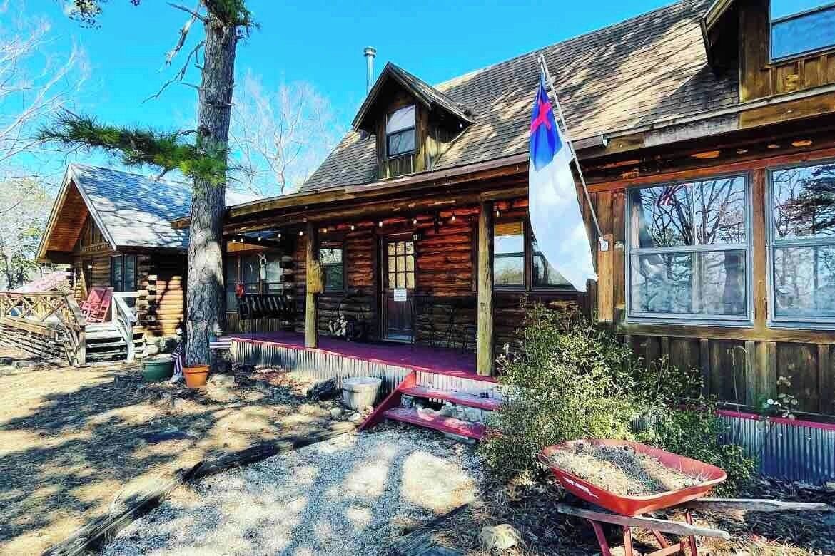 A rustic log cabin with a porch, a flag, and a wheelbarrow in the foreground.